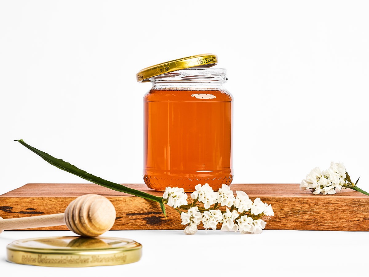 hochwertige Produktfotografie eines Honigglases mit Holzlöffel, kombiniert mit Blüten und natürlichem licht, food styling